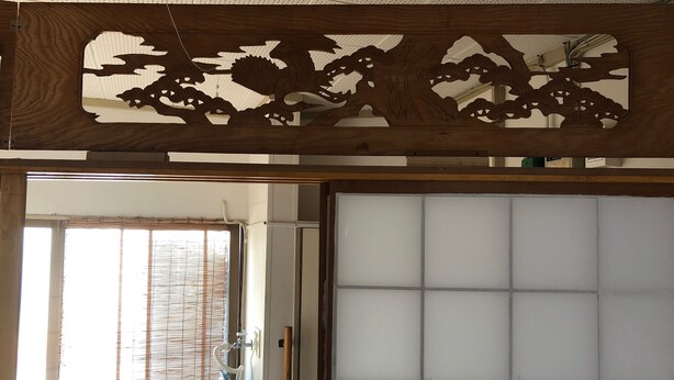 Una habitación con una puerta corrediza de fusuma, un panel decorativo de madera sobre la puerta y una ventana con persianas.