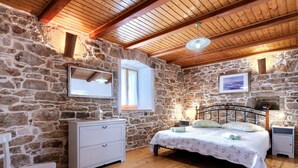 A bedroom with stone walls, wooden ceiling beams, a bed with a floral bedspread, a bedside table, a nightstand, a dresser, and a chair. - Beautiful private villa for 7 people with WIFI, A/C, private pool, TV and patio (Pazin)