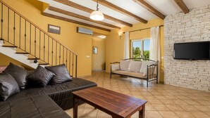 Un salon comprenant un canapé, une table basse en bois, un mur en pierre, un téléviseur fixé au mur et un escalier avec des poutres en bois.