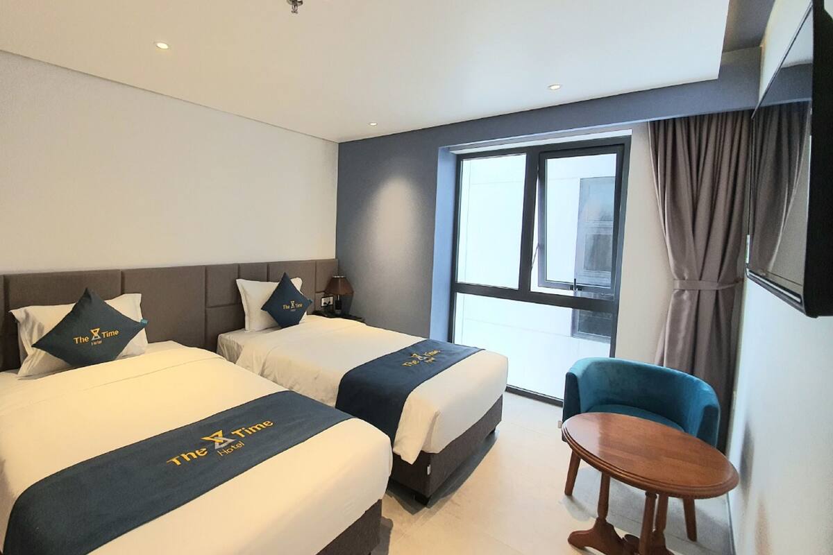 A hotel room with two beds, a small table, a chair, a window with curtains, and a mounted TV.