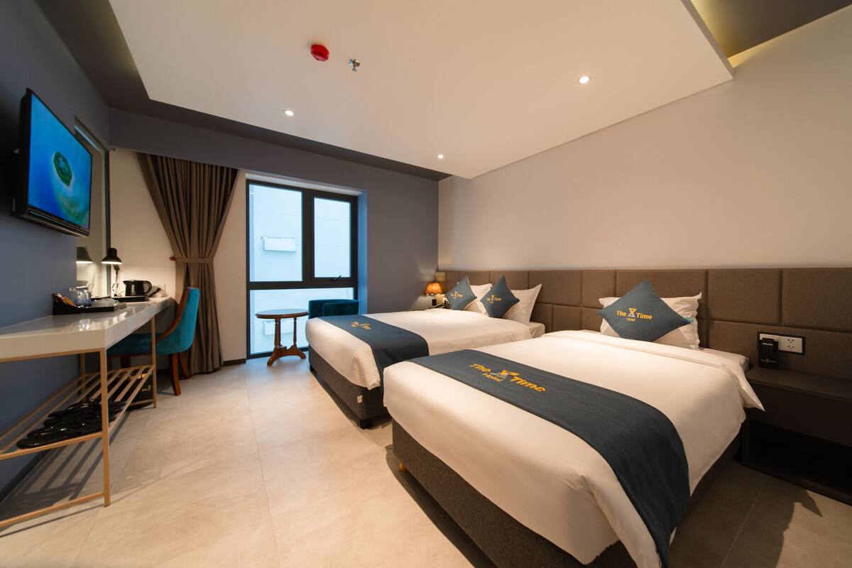 A hotel room with two beds, a desk, a chair, a TV, and a window with curtains.