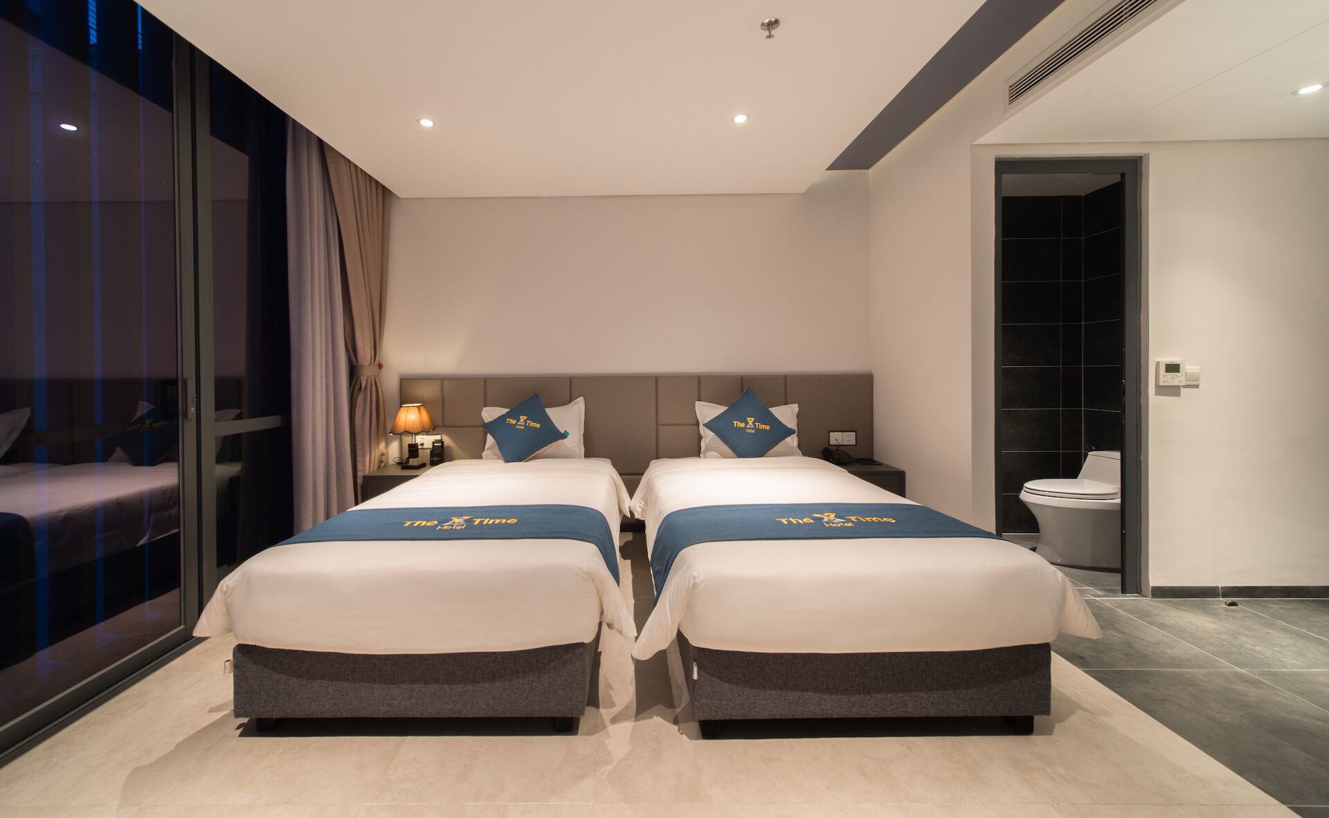 Habitación de hotel con dos camas, una mesita de noche, una lámpara y vistas al exterior.