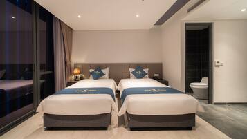 Habitación de hotel con dos camas, una mesita de noche, una lámpara y vistas al exterior.