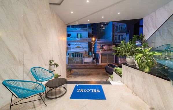 Un vestíbulo de hotel con piso de mármol, una alfombra de bienvenida, sillas azules y plantas en macetas.