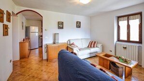 Ein Wohnzimmer mit Sofa, Couchtisch und Esstisch. Es gibt eine Küchenzeile mit Kühlschrank und Mikrowelle. Das Zimmer verfügt über ein Fenster mit Vorhängen und eine Heizung.