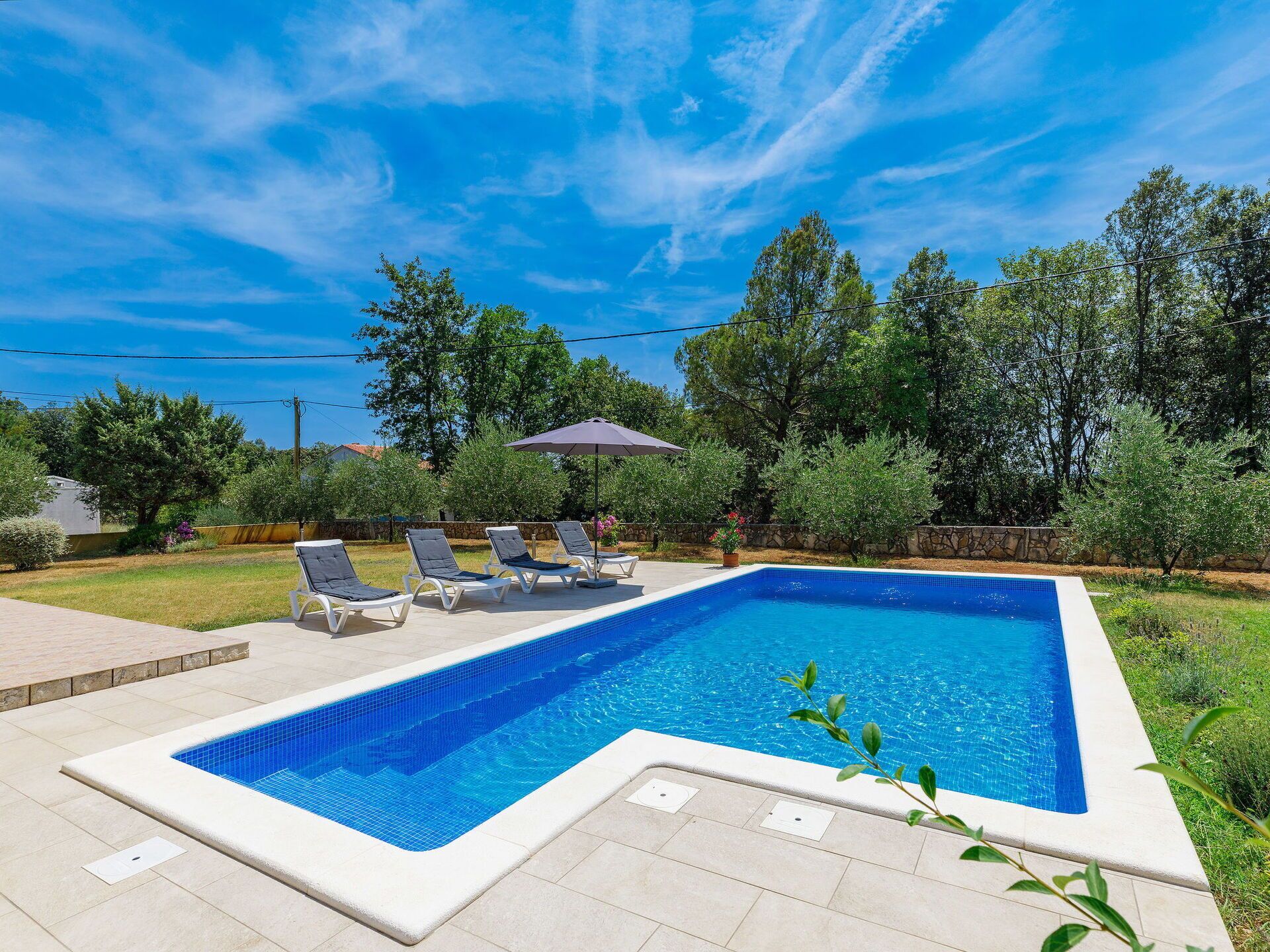 A rectangular swimming pool with a tiled edge, surrounded by a paved deck. There are four lounge chairs and a single umbrella.