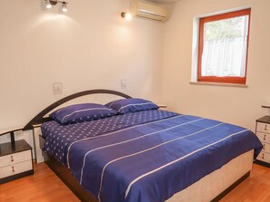 Une chambre à coucher comprenant un lit, une table de chevet, une commode, un appareil de climatisation et une fenêtre donnant sur des arbres.
