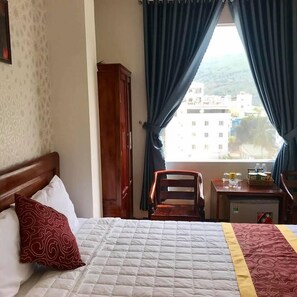 A hotel room with a bed, a wooden chair, a small table with a bottle and a box, and a view of buildings through the window.
