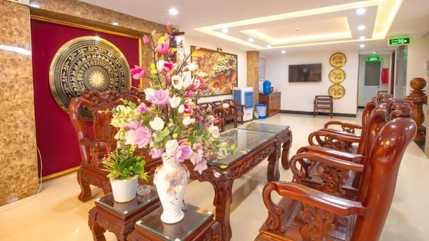 A hotel lobby with a reception desk, wooden chairs, a large decorative vase with flowers, and a wall with a circular emblem. - New Day Hotel Quy Nhon (Quy Nhon Nam)