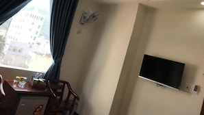 A hotel room with a large window, a flat-screen TV mounted on the wall, a ceiling fan, and a small table with a chair. - New Day Hotel Quy Nhon (Quy Nhon Nam)