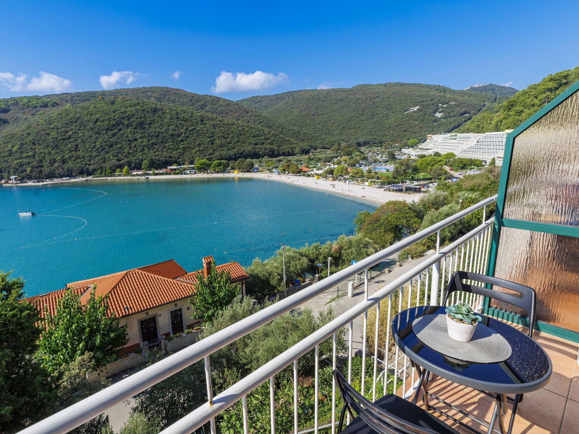 A balcony with a table and chair overlooking a coastal area with a beach and hills.