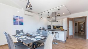 Un'area pranzo moderna con un tavolo rettangolare apparecchiato per quattro persone, sedie rivestite in grigio, illuminazione a sospensione e una cucina sullo sfondo.