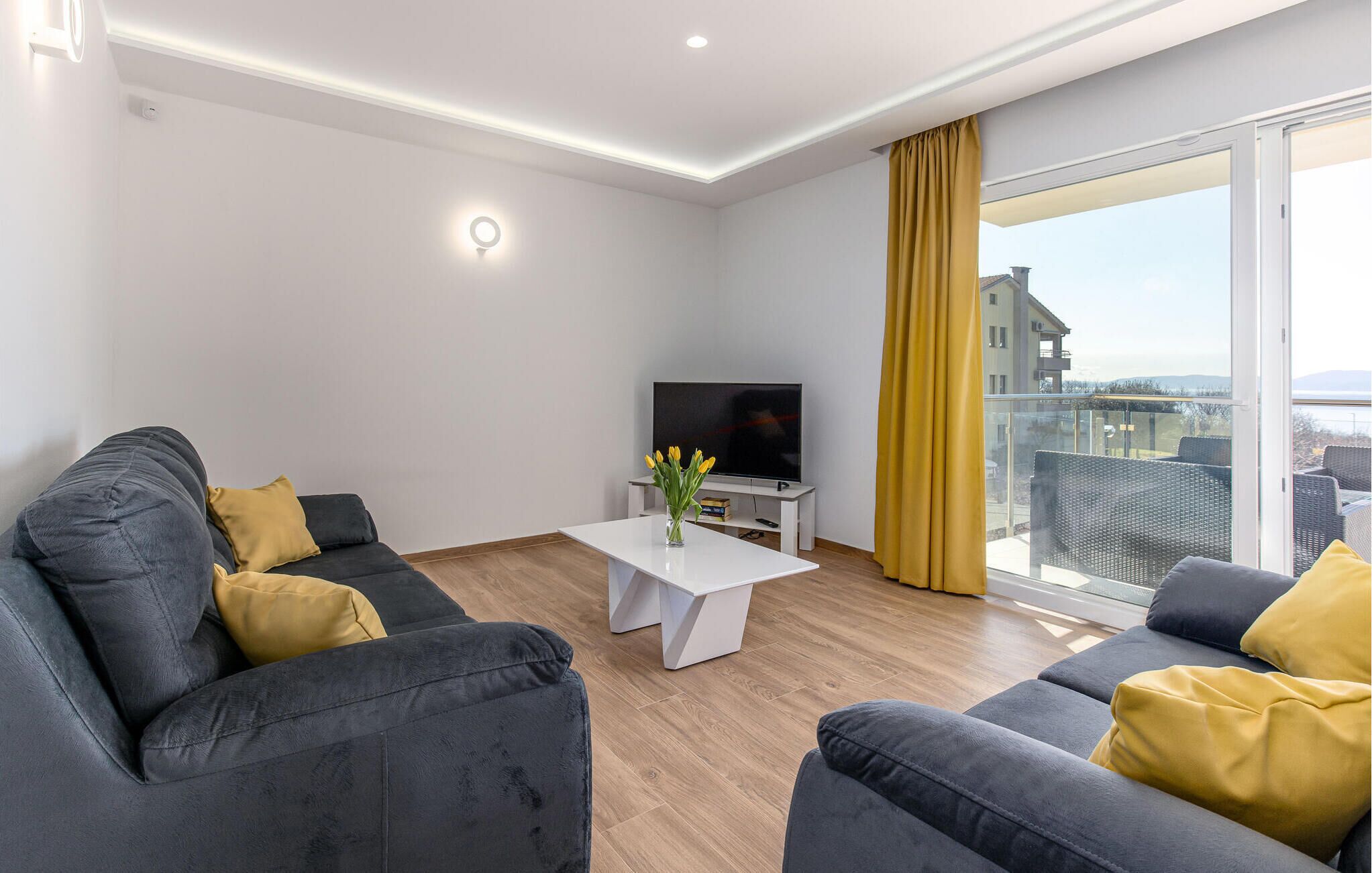 A modern living room with a grey sofa set, a white coffee table with a vase of flowers, and a TV on a white stand.