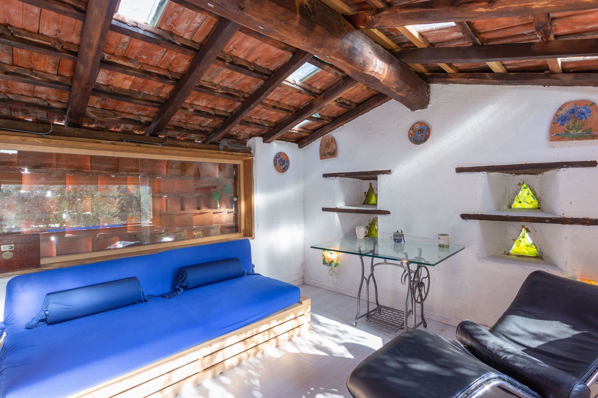 Una habitación con techo de madera, un sofá azul, una mesa de vidrio y estantes con adornos.