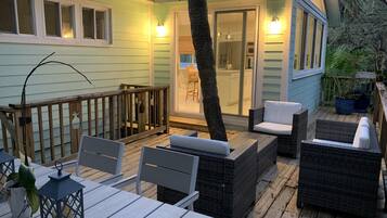 Une terrasse en bois avec des meubles d’extérieur, une porte vitrée donnant sur une cuisine et une plante en pot.