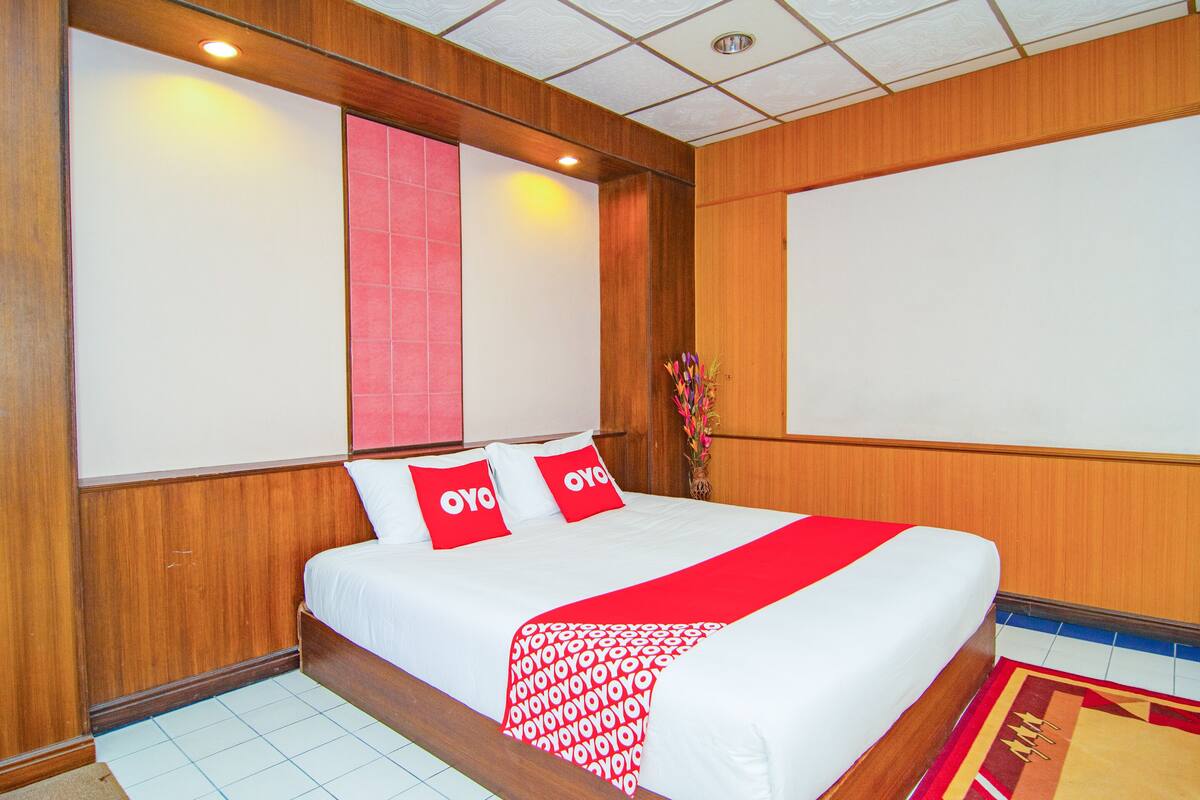 Une chambre d’hôtel avec un grand lit, des boiseries, une couvre-lit rouge et blanche, et un oreiller rouge et blanc orné du logo de l’hôtel.