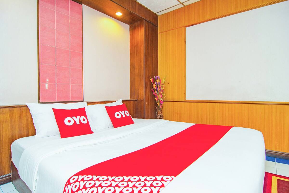 Une chambre d’hôtel avec un lit, deux oreillers rouges ornés du texte « OYO », une couvre-lit rouge et blanche et une tête de lit en bois.