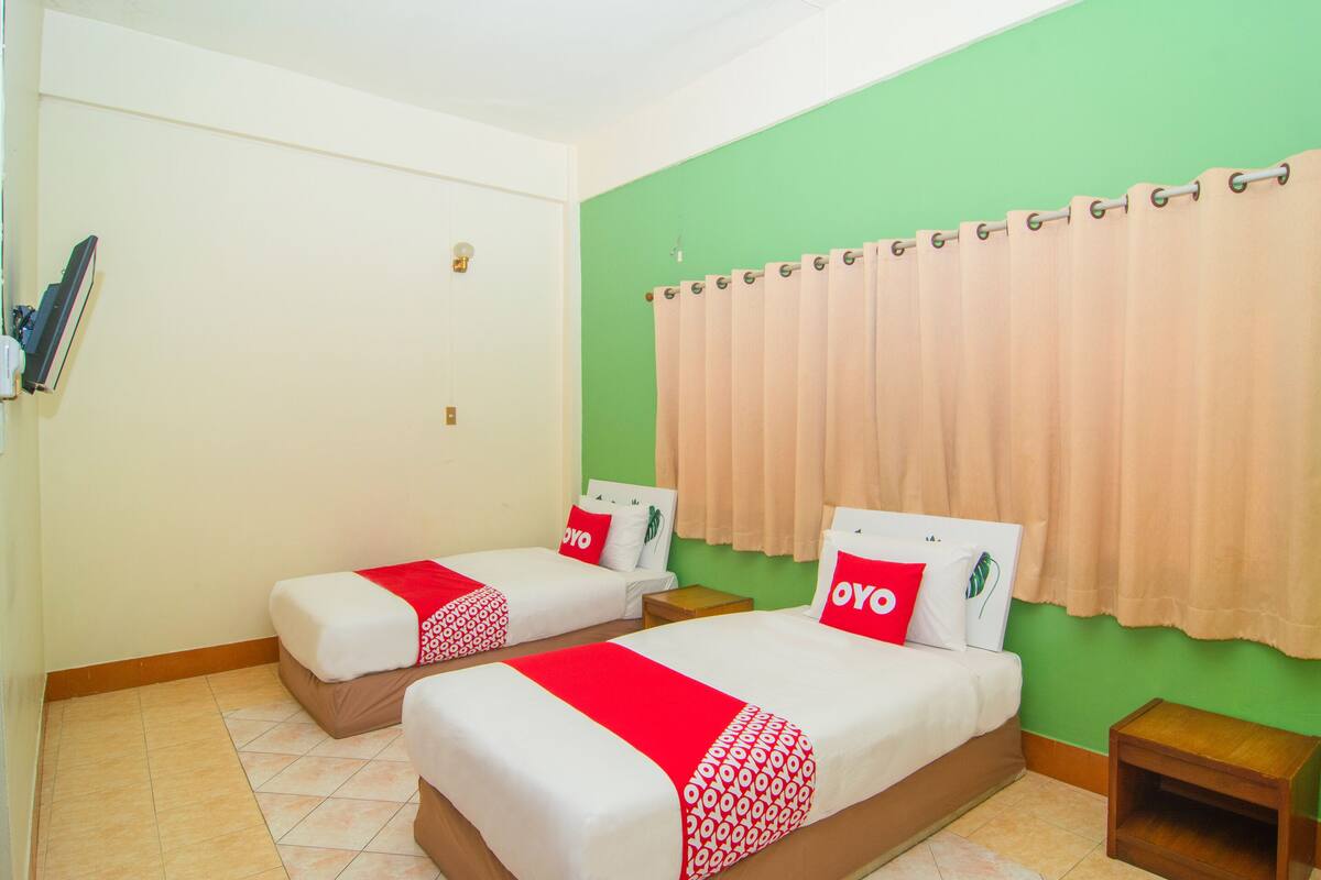 Une chambre d’hôtel avec deux lits, chacun recouvert d’une courtepointe rouge et blanche, une petite table de chevet en bois et un mur d’accent vert.