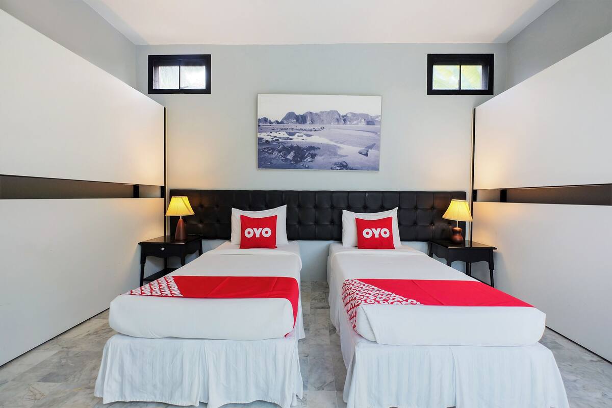 A hotel room with two beds, each with a red pillow labeled "OYO," a large framed picture on the wall, and two bedside tables with lamps.
