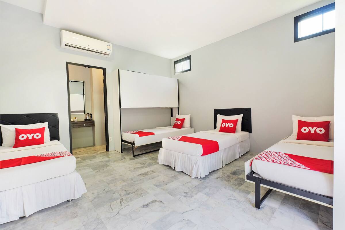 A hotel room with three beds, each with white bedding and red pillows labeled "OYO". There is a large mirror on the wall, an air conditioning unit above the beds, and a small window on the right wall.