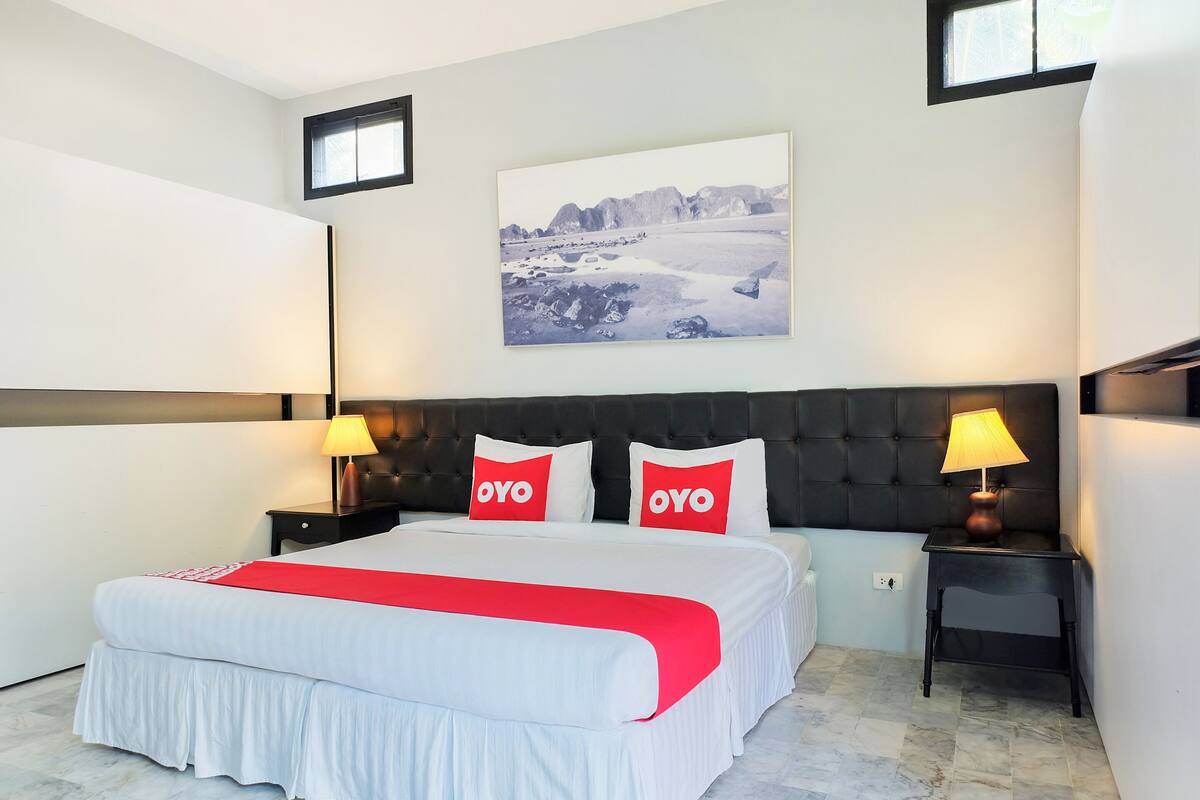 A modern hotel room with a large bed, two bedside tables with lamps, a headboard, a framed picture on the wall, and a window.