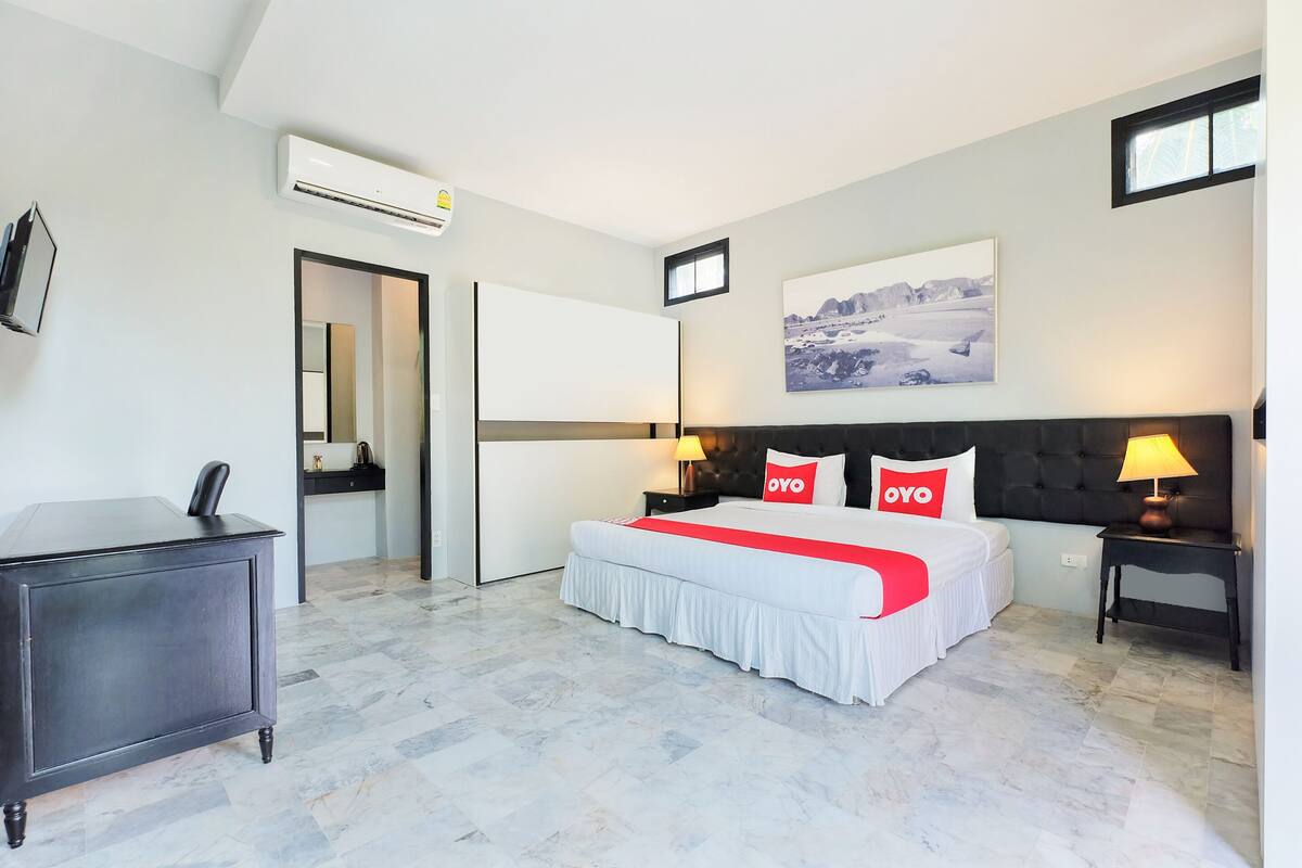 A modern hotel room with a large bed, a bedside table with a lamp, a wardrobe, and a painting on the wall.
