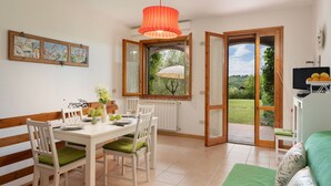 Uma área de refeições com mesa branca, cadeiras acolchoadas verdes, luminária laranja pendente e janela com vista para vegetação.