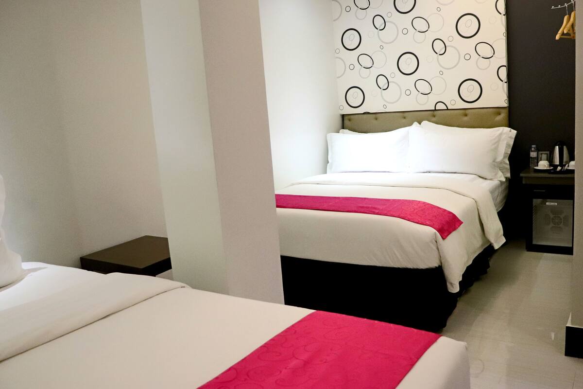 Une chambre d’hôtel avec deux lits, une tête de lit ornée d’un motif circulaire, une petite table de chevet et une table de nuit avec une bouilloire.