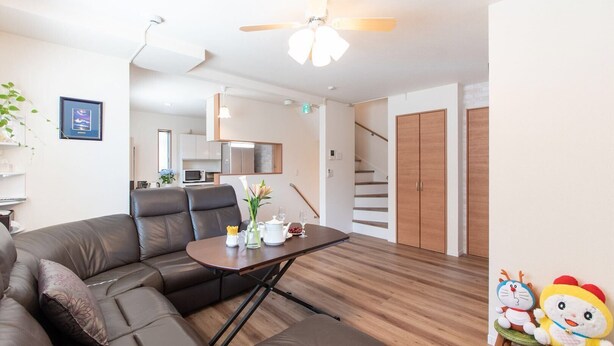 A modern living room with a sectional sofa, a dining table with a vase of flowers, a kitchen in the background, and a staircase leading to a wooden door. - Mori House In Nishinippori (Tokyo)