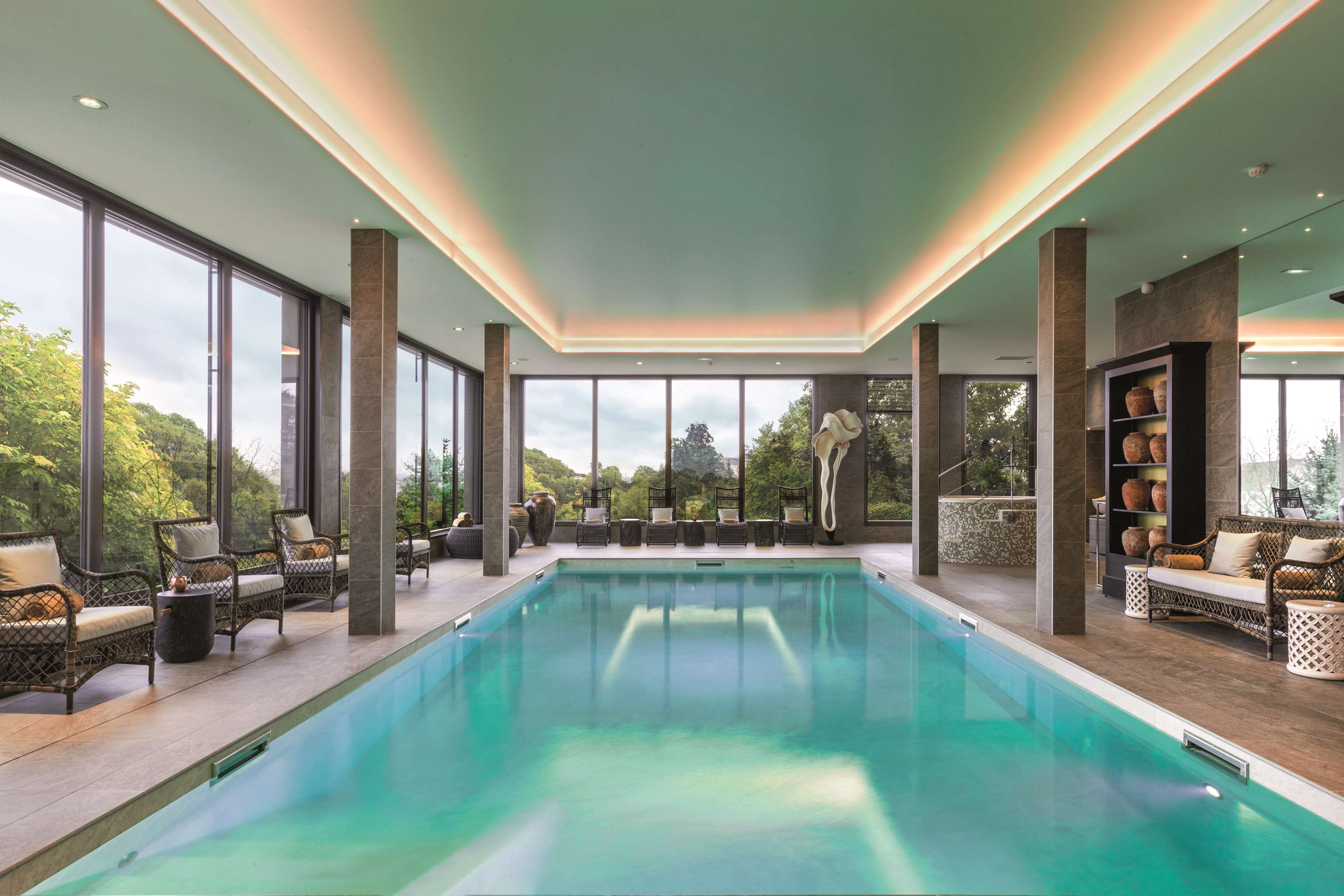 Una piscina interior moderna con amplios ventanales, mobiliario de mimbre y vistas al paisaje circundante.