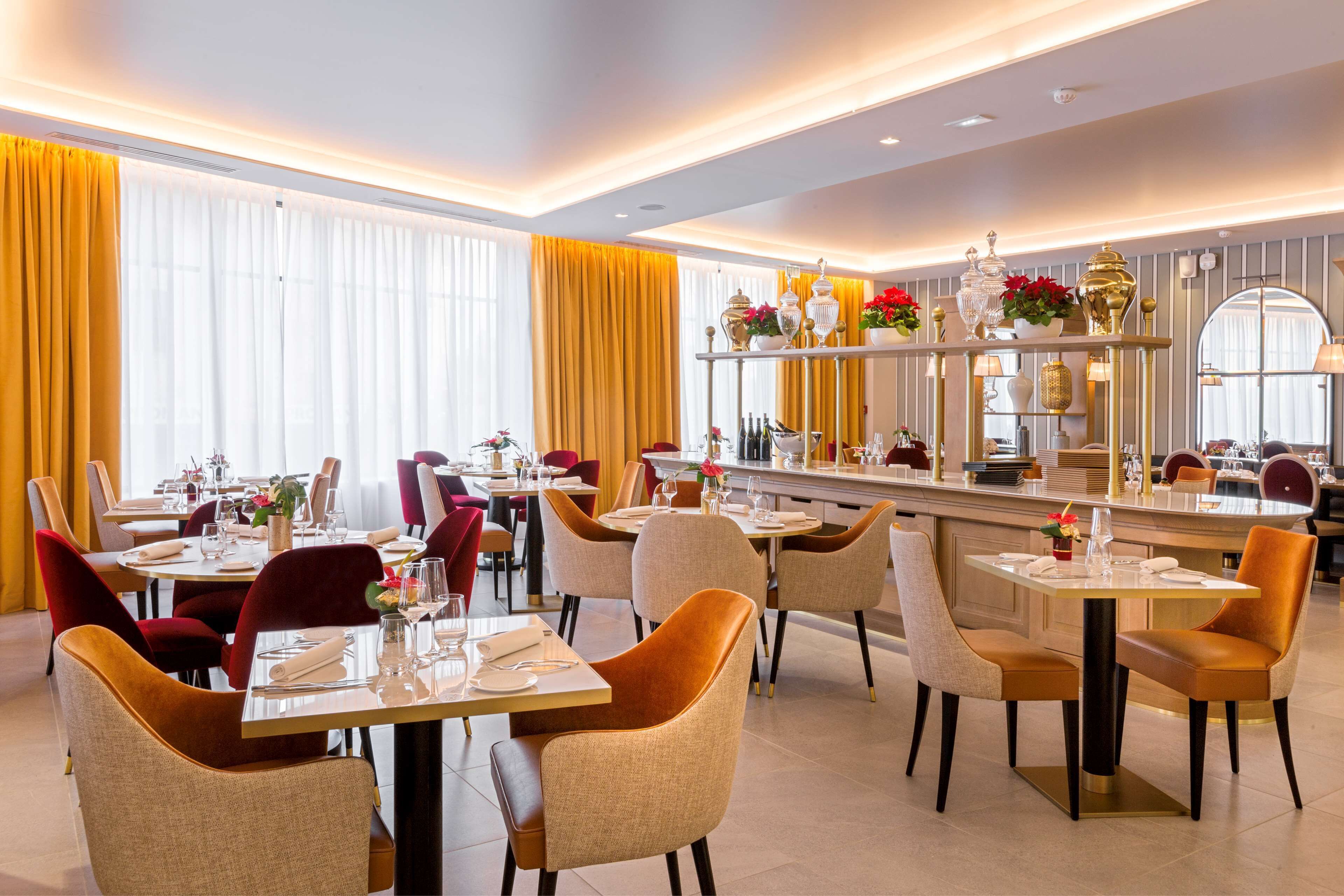 Un comedor bien iluminado con varias mesas y sillas, una zona de bar con mostrador, y amplias ventanas con cortinas.