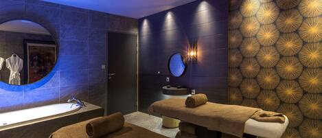 Un spa moderno con una bañera grande, un espejo redondo, una camilla de masaje y una pared con un diseño geométrico.
