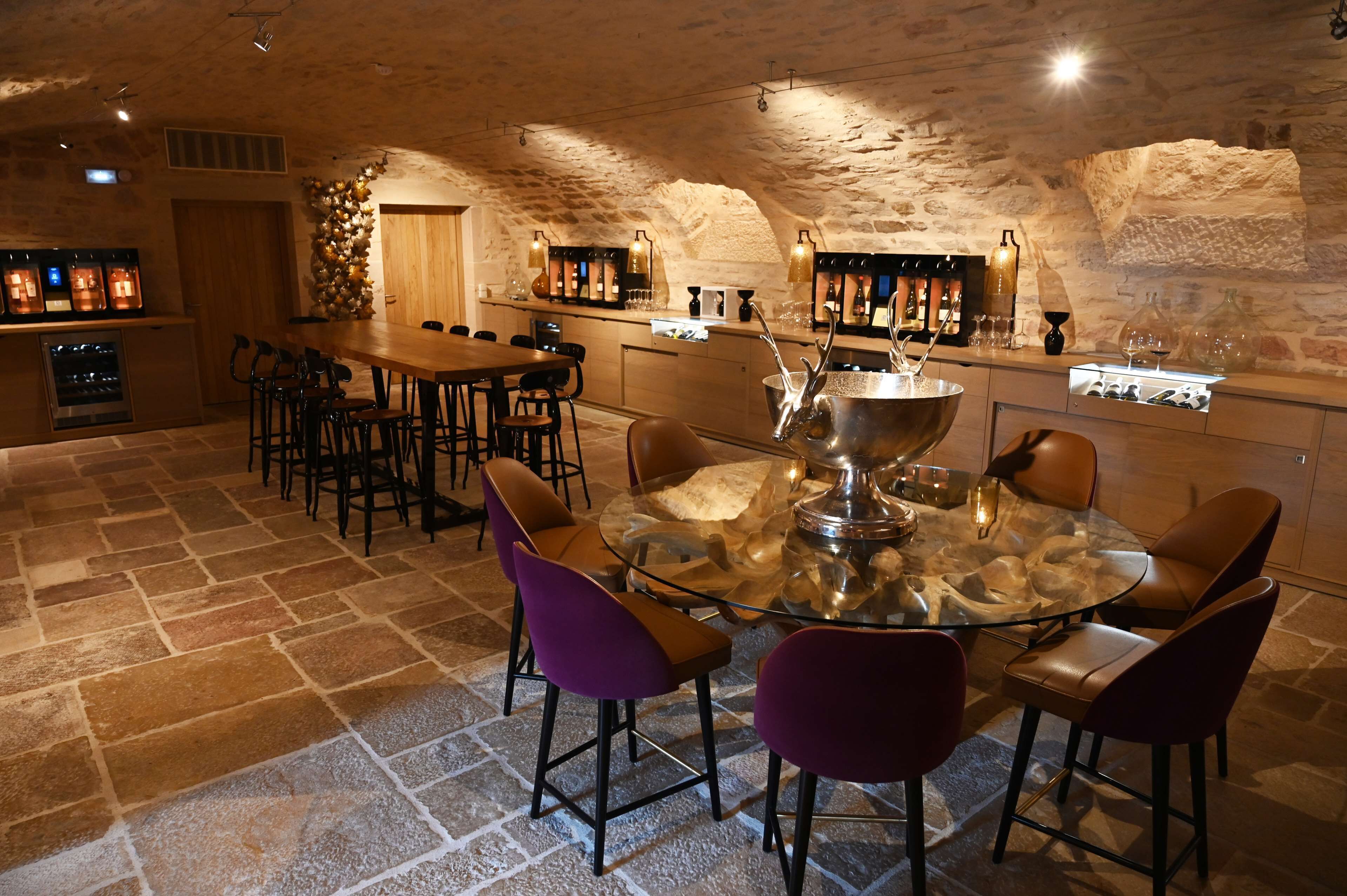 A stone-walled bar area with a round glass table and leather chairs, equipped with a large metal bowl and bar tools.