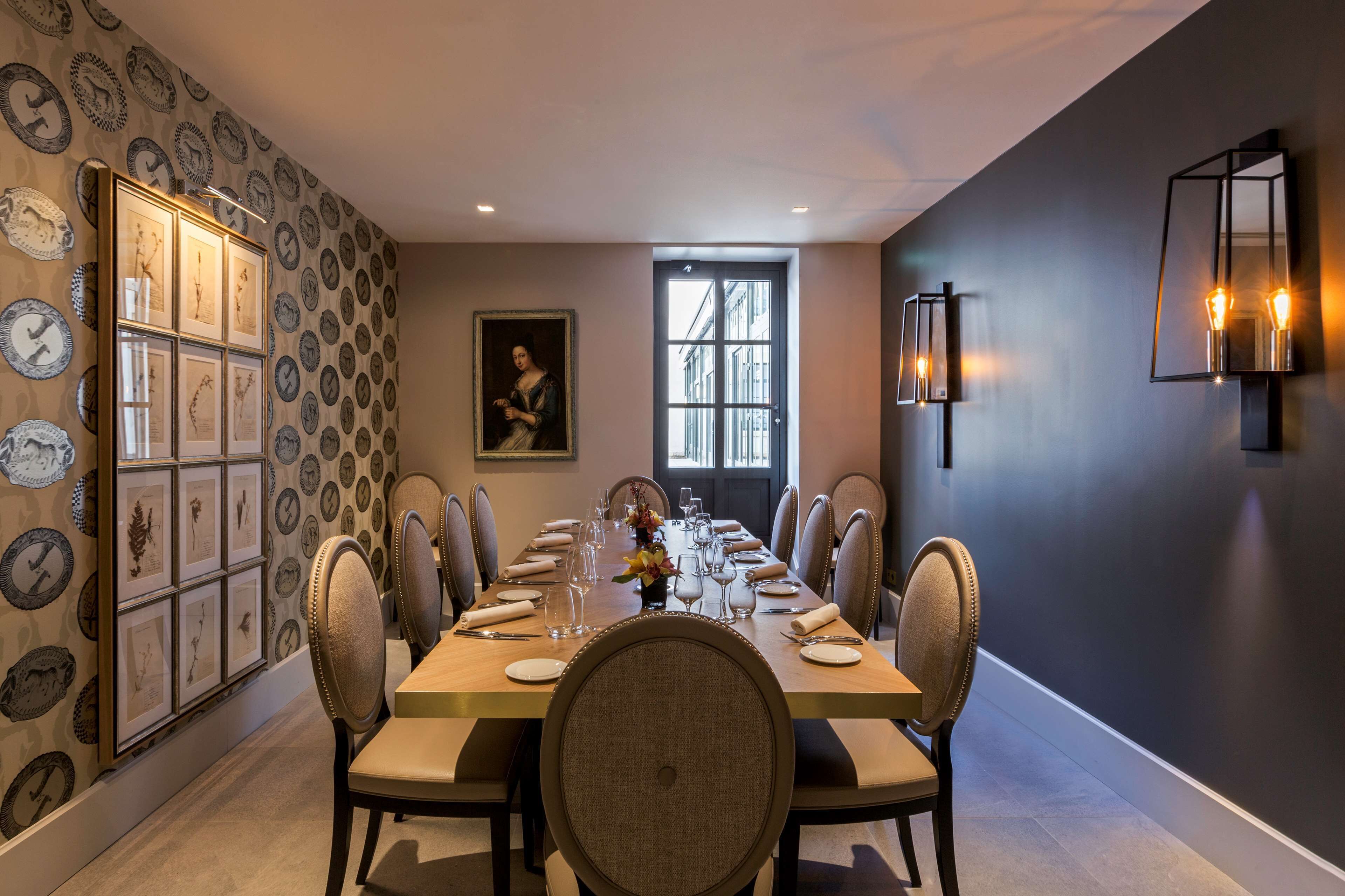 Un comedor con una mesa larga dispuesta para una comida, rodeada de sillas. Hay una obra de arte enmarcada en la pared y una ventana que deja entrar luz natural.