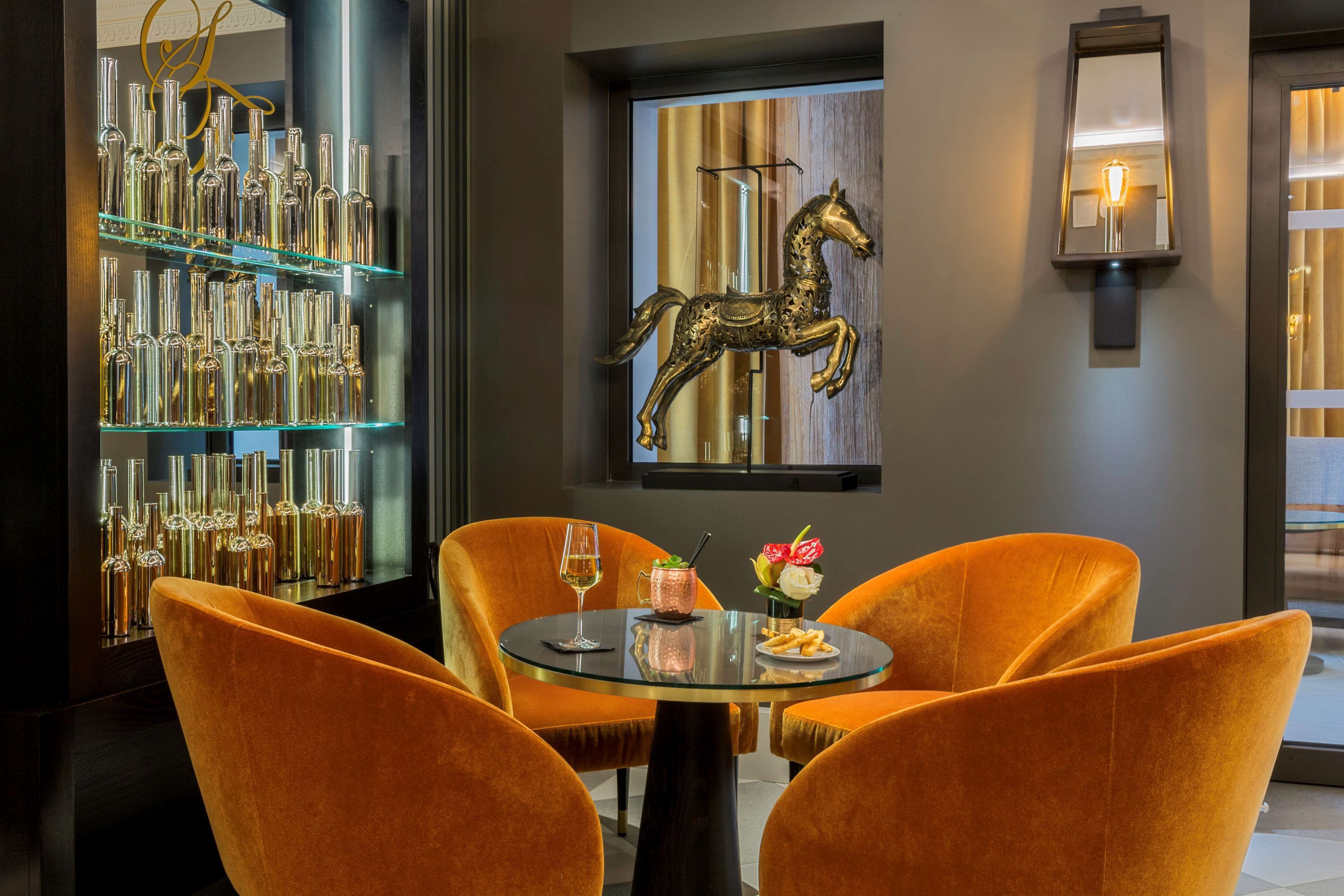 A dining area with a glass table, orange upholstered chairs, a decorative horse sculpture, and a glass display of bottles.