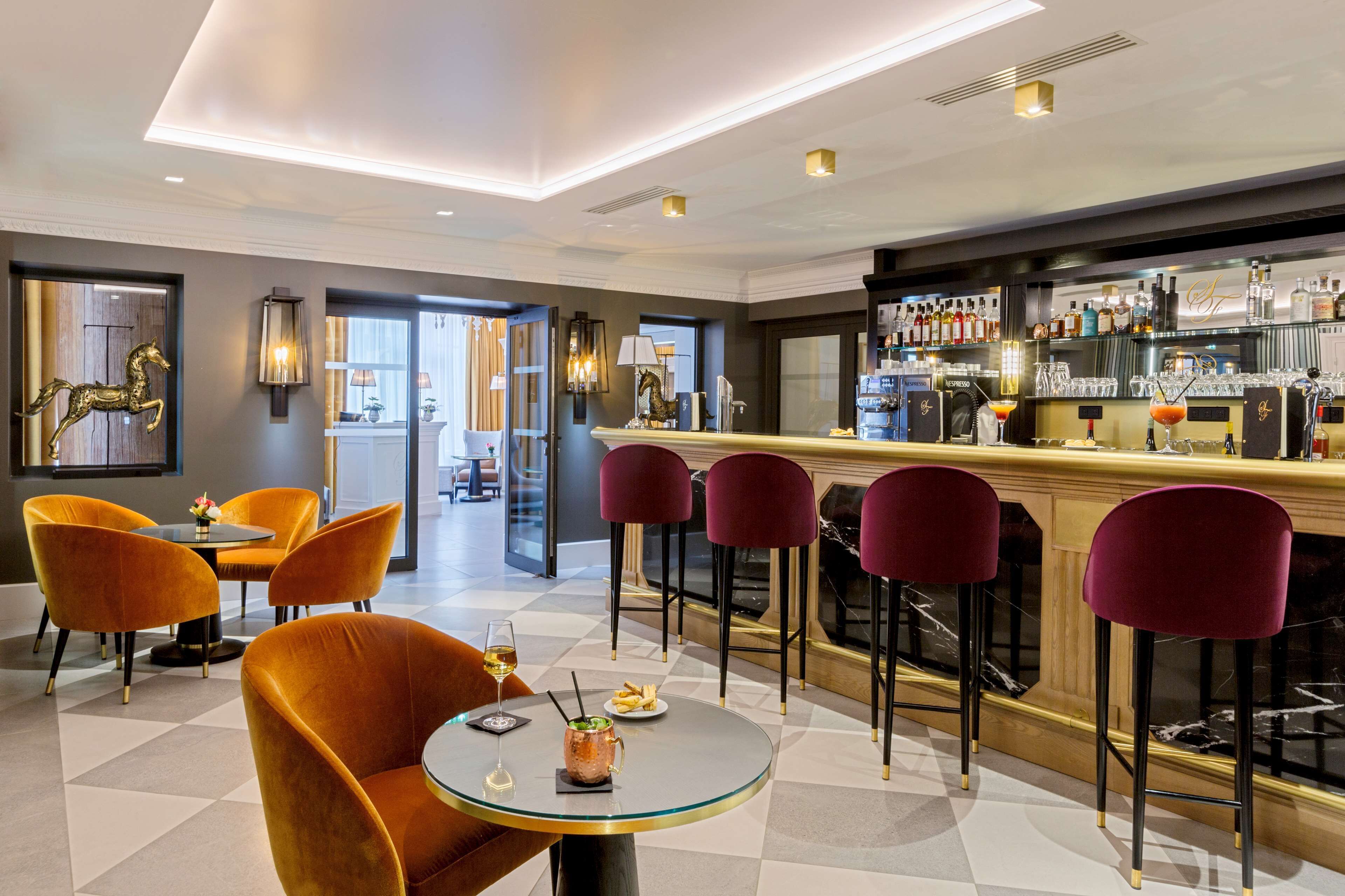 Een moderne bar met een marmeren aanrecht, diverse zitmogelijkheden zoals ronde tafels en barstoelen, en een selectie flessen en glazen op de planken.