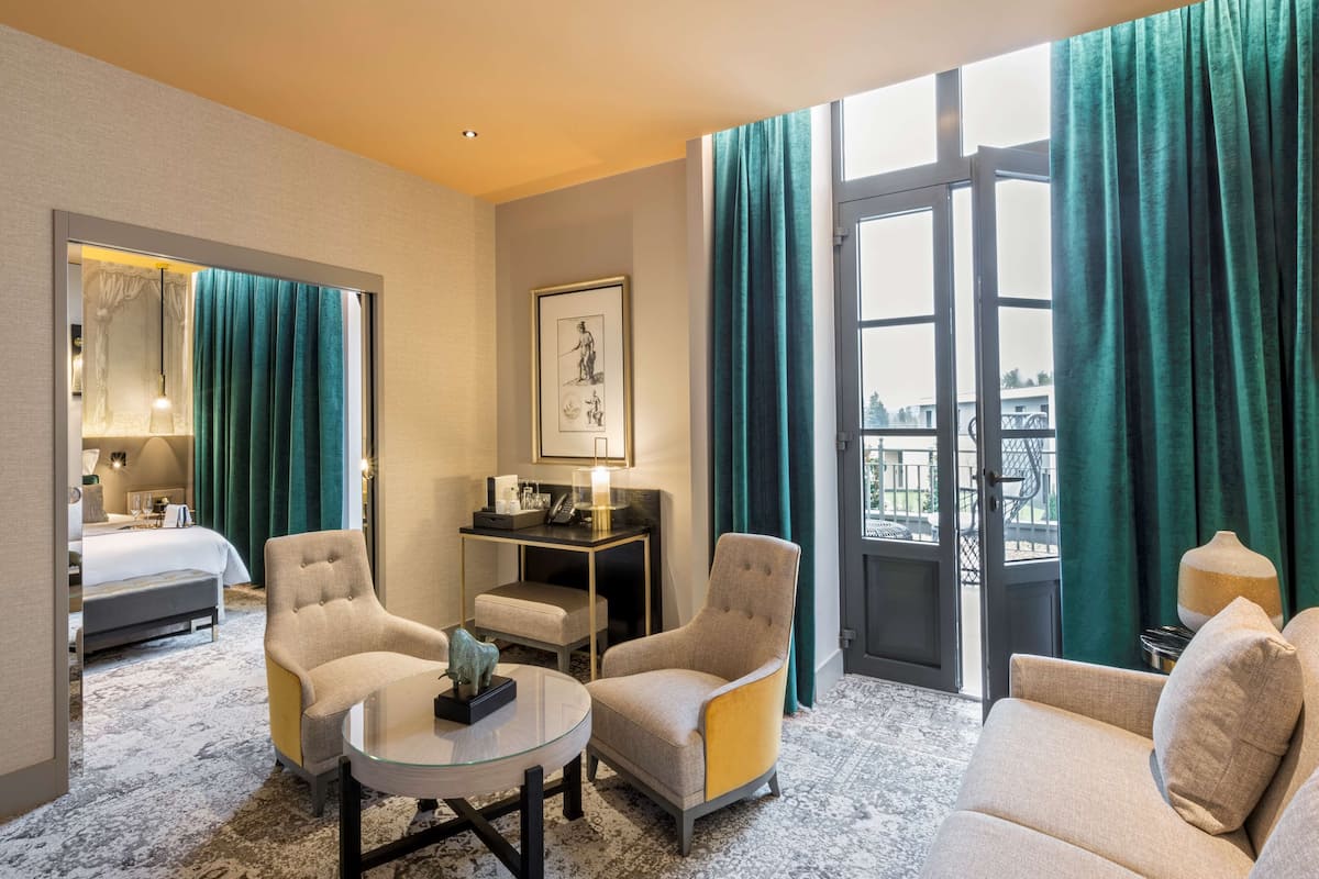 A hotel room with a bed, two armchairs, a small table, a desk with a lamp, and a large window with curtains.
