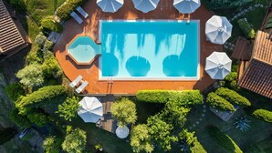 Une vue aérienne d’une piscine rectangulaire avec un jacuzzi hexagonal, entourée de parasols blancs et de chaises longues, le tout aménagé dans un jardin paysager avec des arbres et une terrasse en briques.
