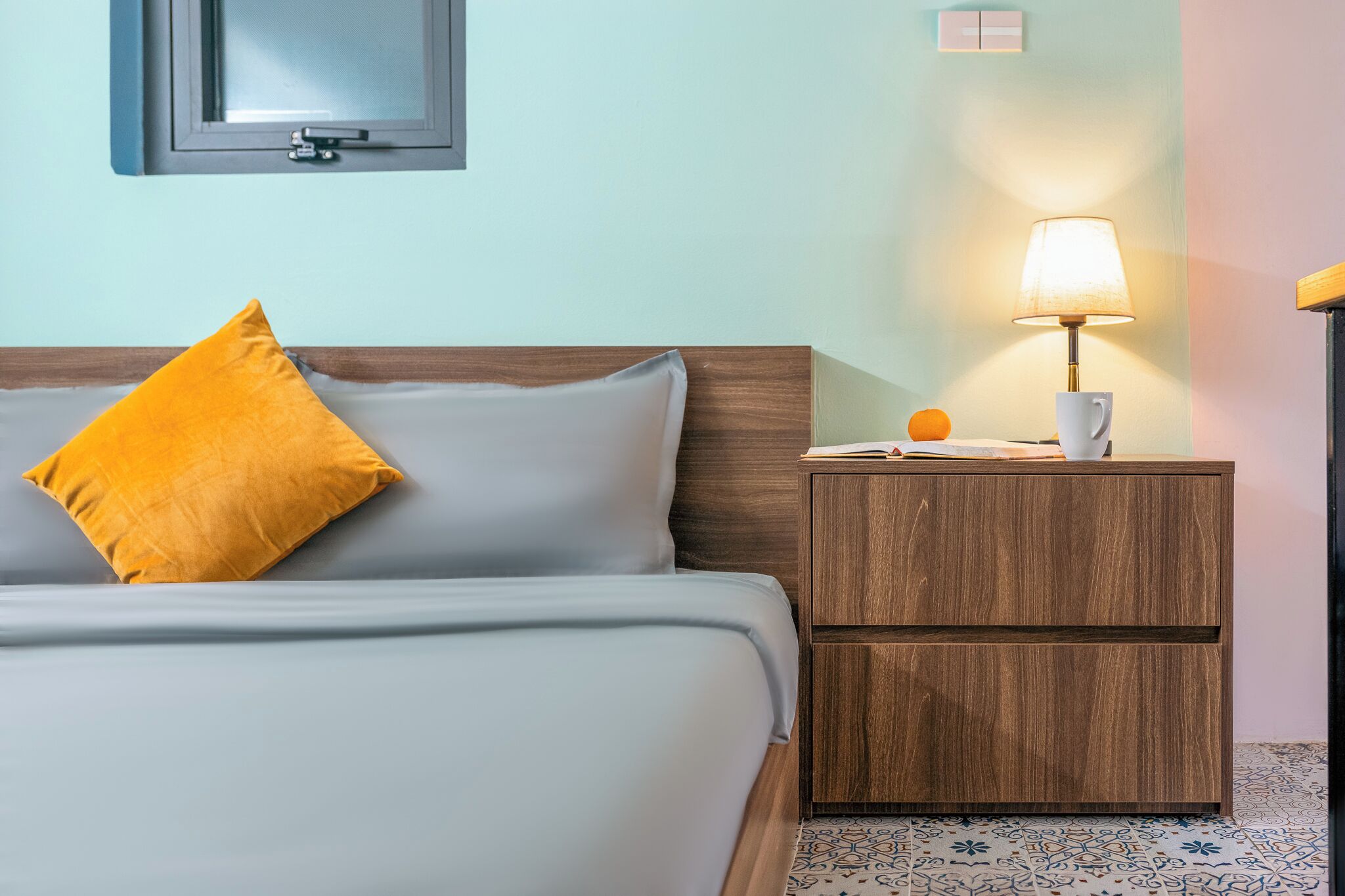 Una habitación de hotel con una cama, una mesita de noche con una lámpara, una naranja y una taza.