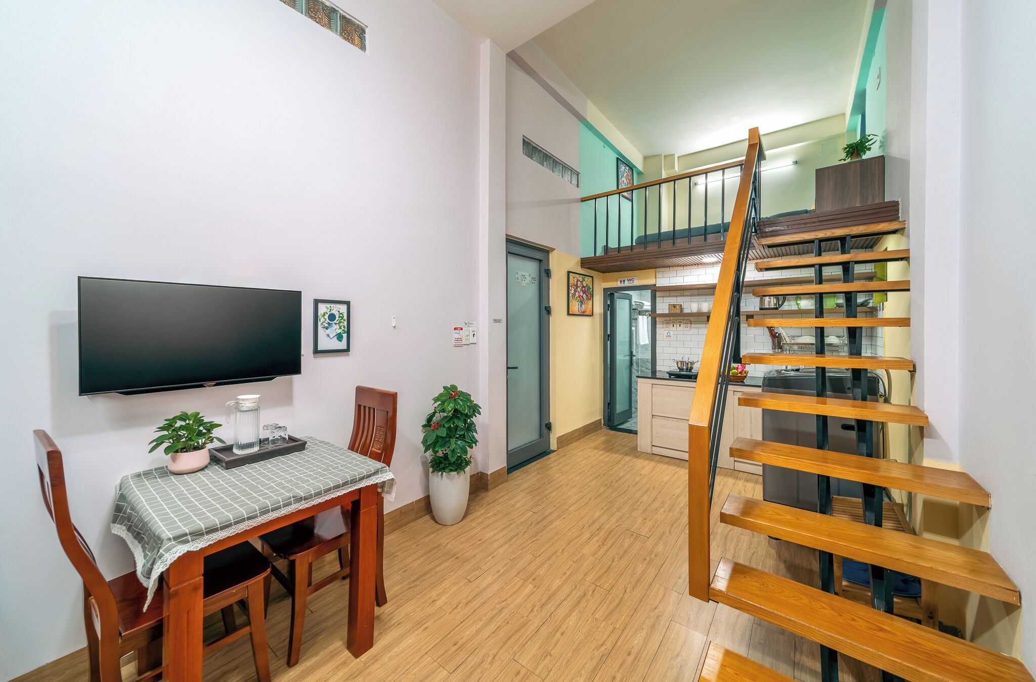 A modern living room with a flat-screen TV, a dining table, chairs, and a staircase leading to an upper level.