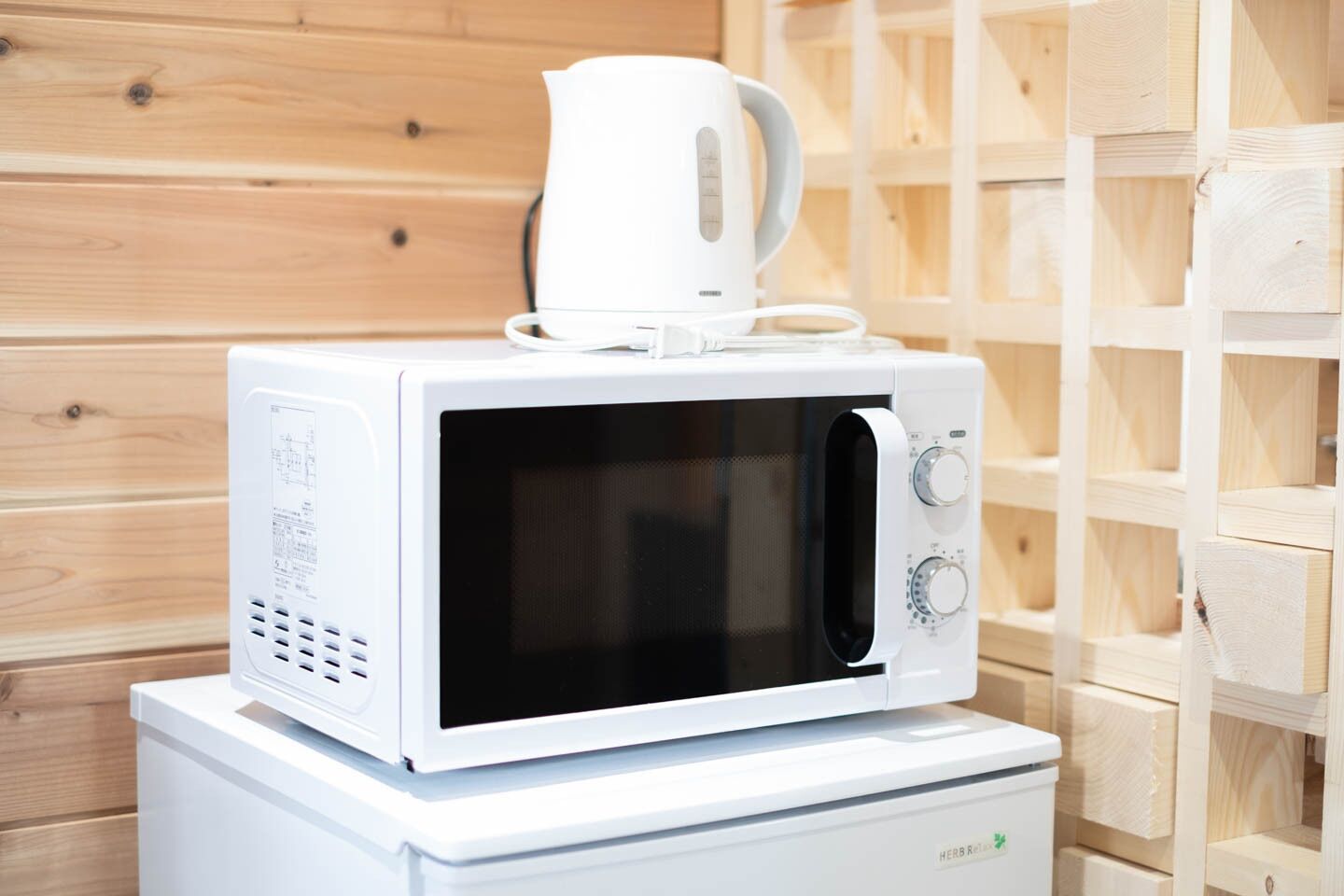 A white microwave on a white appliance, with a white kettle on top, against a wooden wall background.