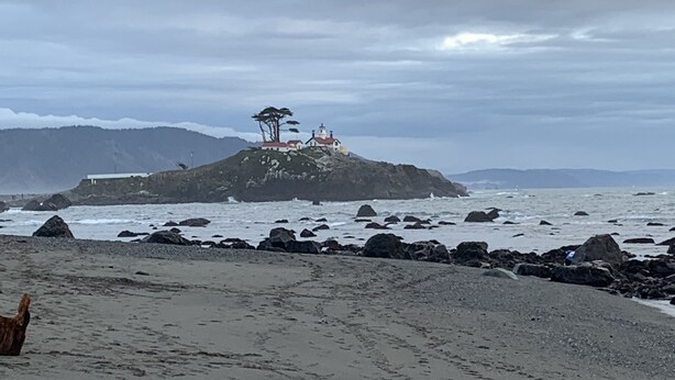 Ein Strand mit felsigem Ufer, ein kleines Gebäude mit rotem Dach und ein einzelner Baum auf einem Hügel.