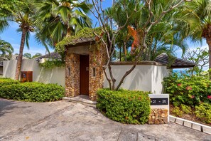 Uma entrada de pedra com porta de madeira, rodeada por vegetação exuberante e palmeiras.