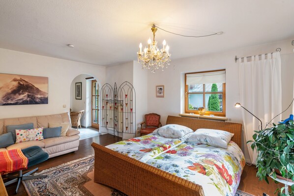 A bedroom with a bed, a sofa, a chandelier, a window with curtains, and a plant.