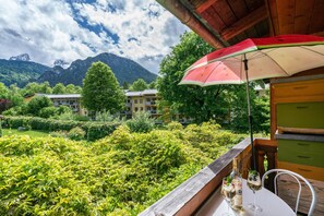 A balcony with a view of lush greenery and mountains, a table with drinks, and a red and white umbrella.