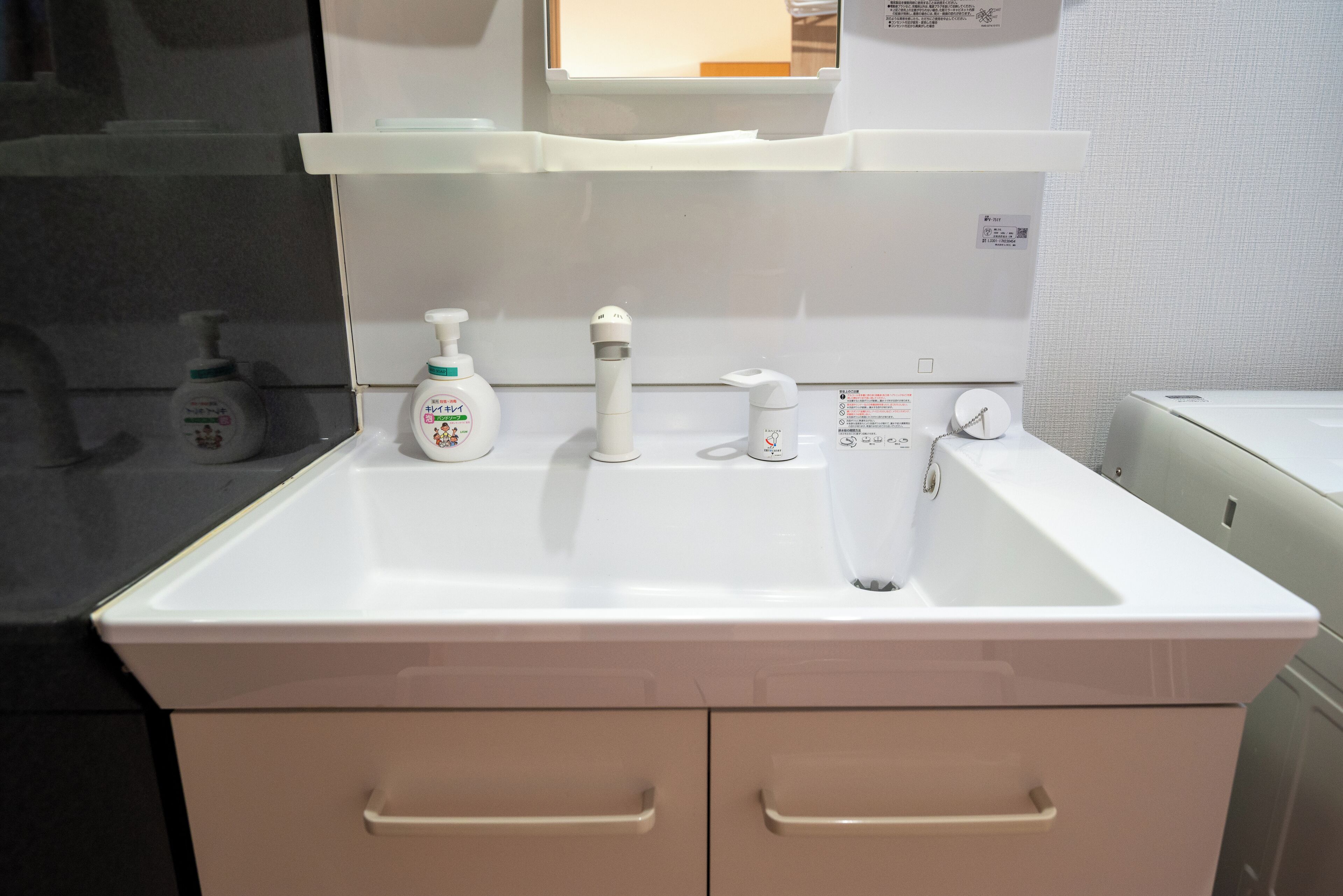 Un lavandino da bagno bianco con due cassetti, uno specchio sopra e tre flaconi di gel disinfettante sul piano.
