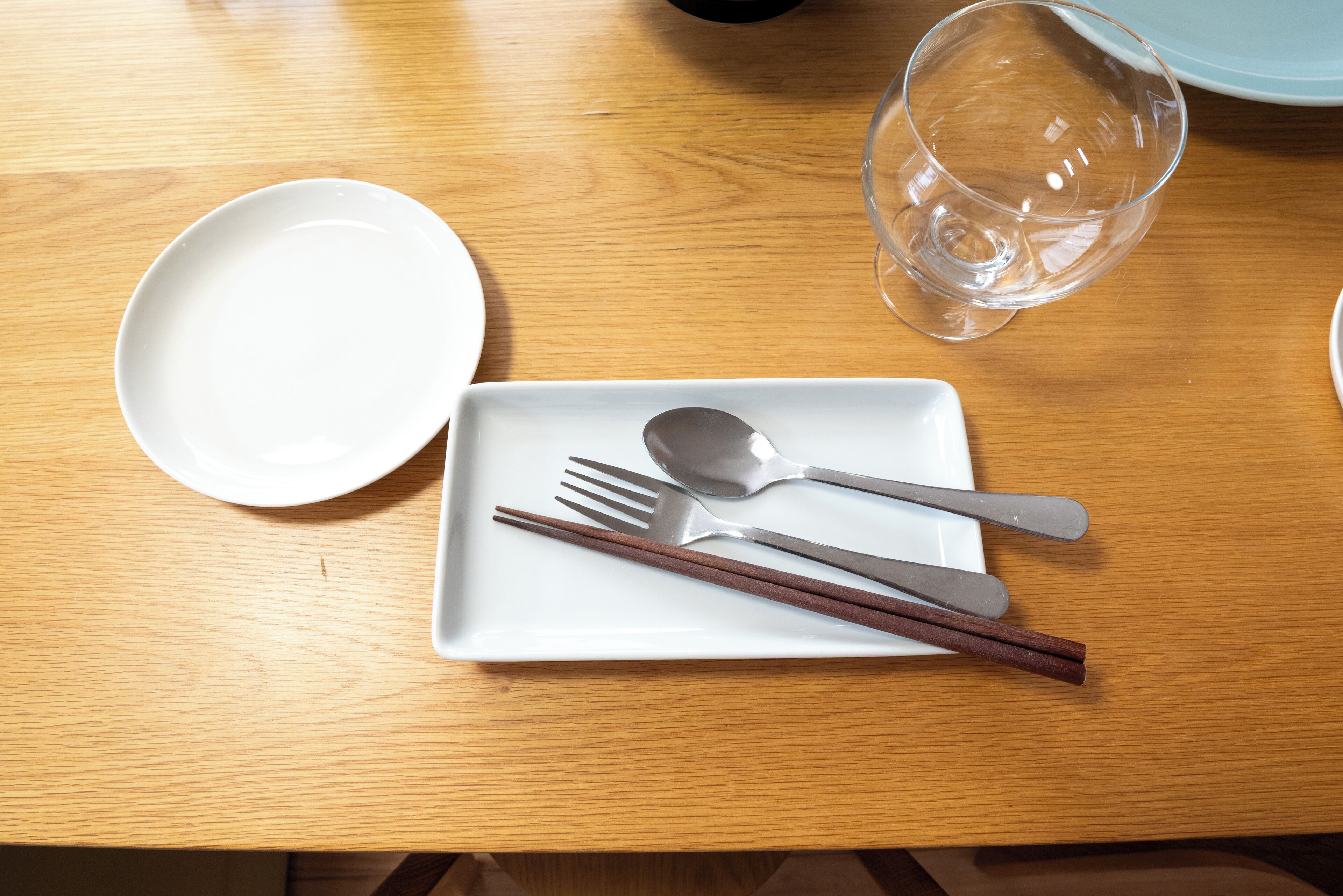 A wooden table with a white plate, chopsticks, a spoon, and a glass.