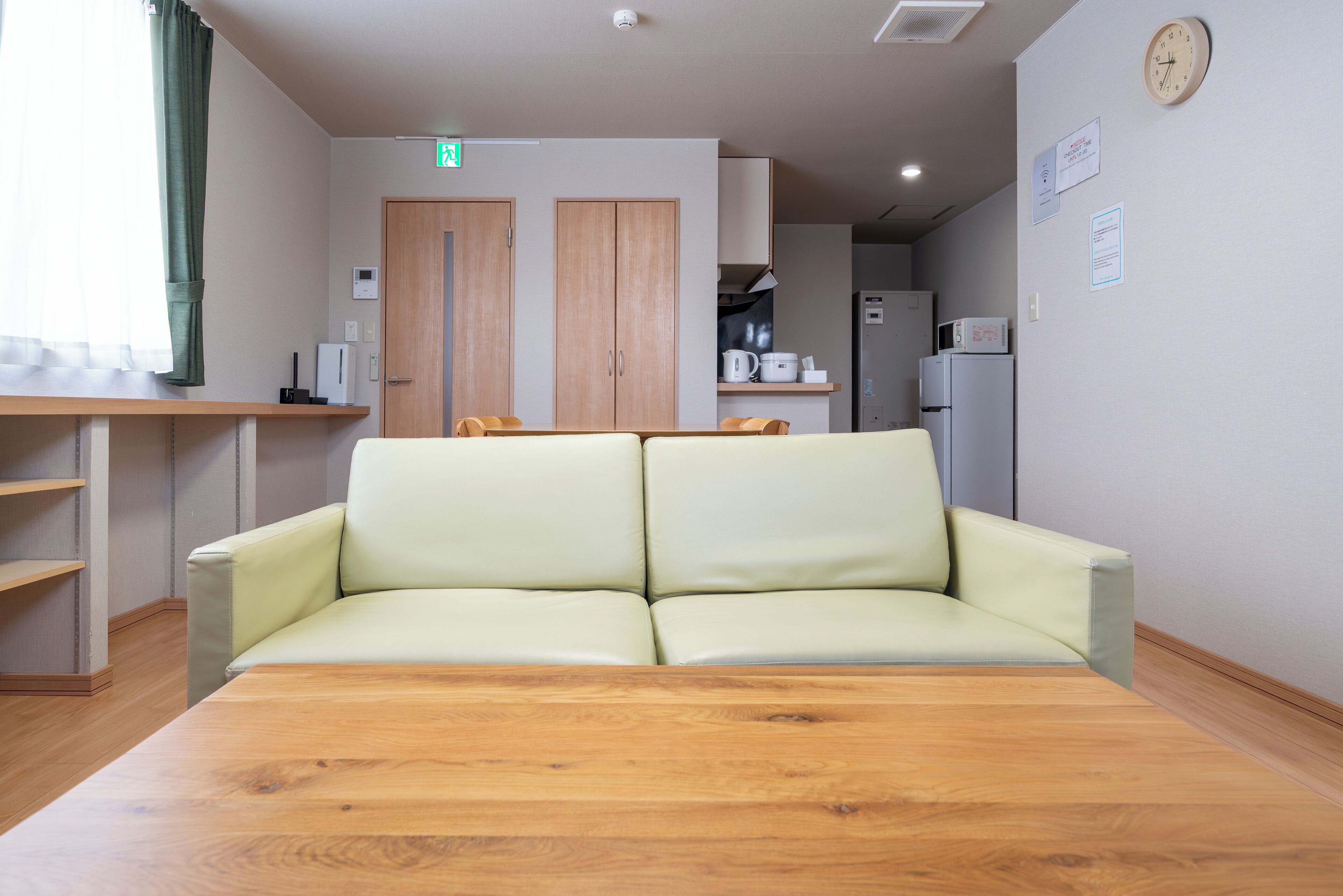 A room with a wooden table, a light-colored sofa, a kitchen area with cabinets, a microwave, and a refrigerator.