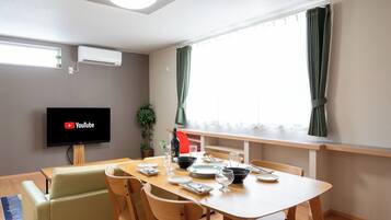 A modern dining area with a wooden table set for four, a flat-screen TV mounted on the wall, and a window with curtains.