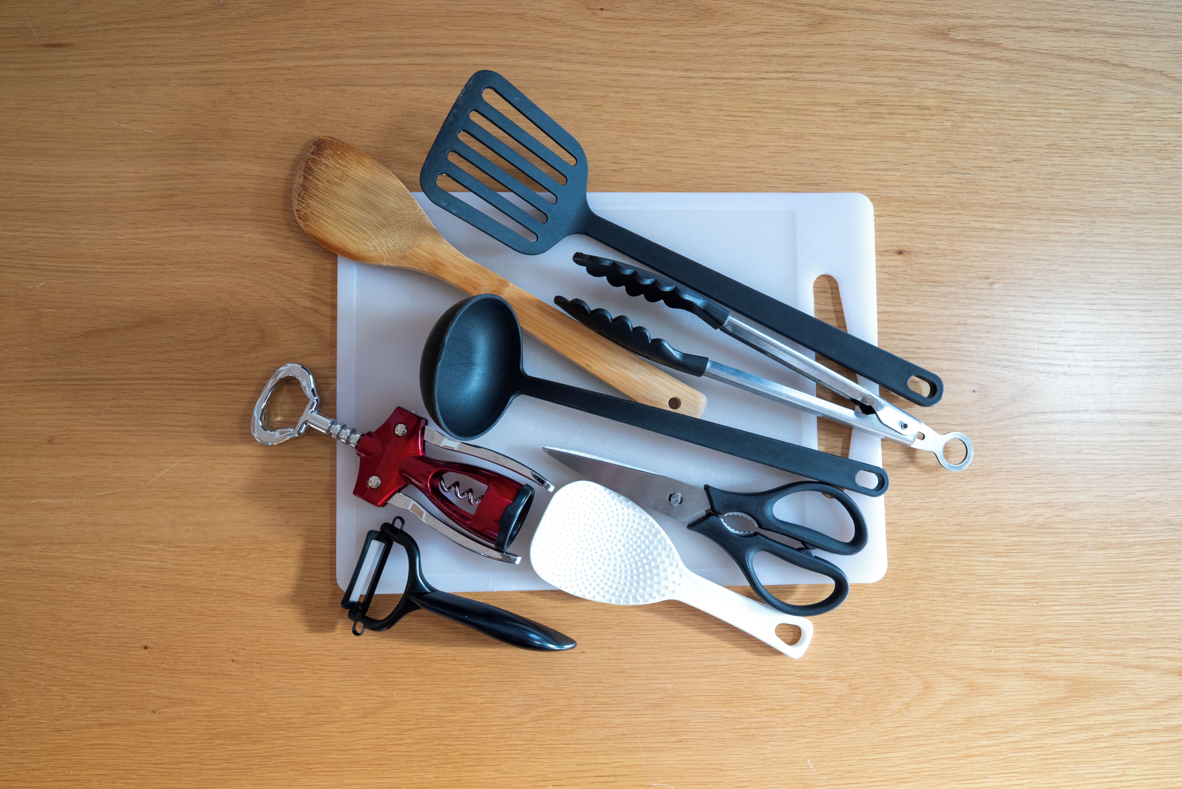 A wooden table with various kitchen utensils, including a wooden spatula, a black spatula, a pair of scissors, a red can opener, and a white grater.